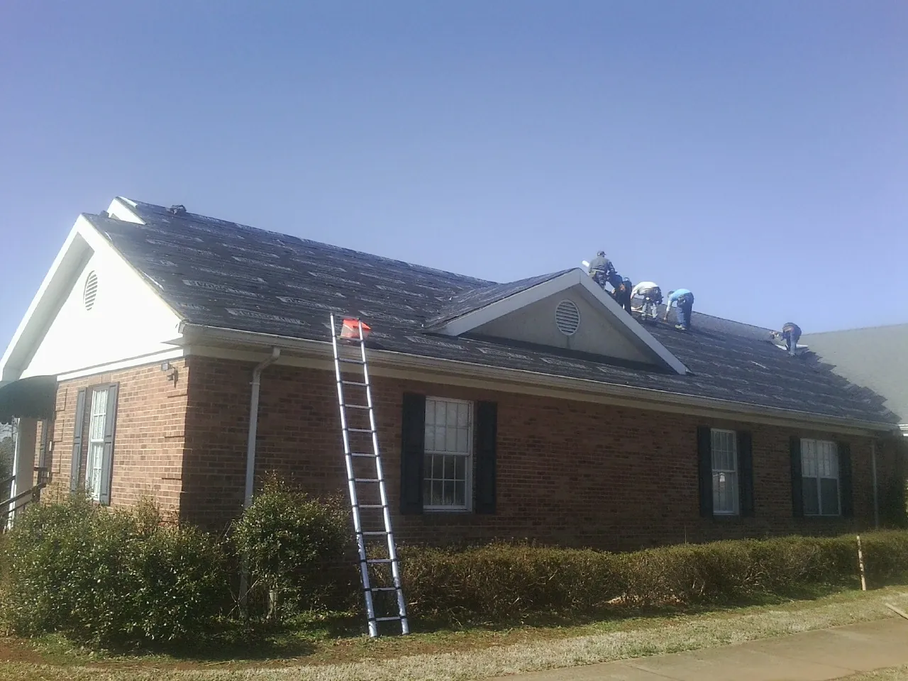 Crew tearing shingles off a brick ranch roof with a ladder set — Metal Roof Repair work by our crew in Saint Johnsville