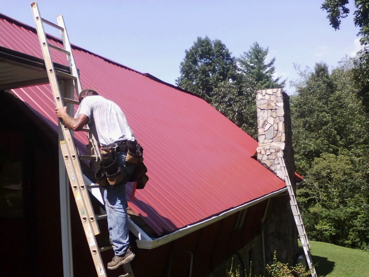 Roofer on a ladder beside a red metal roof and stone chimney — Soffit & Fascia Repair by Roofing Brothers in Saint Johnsville